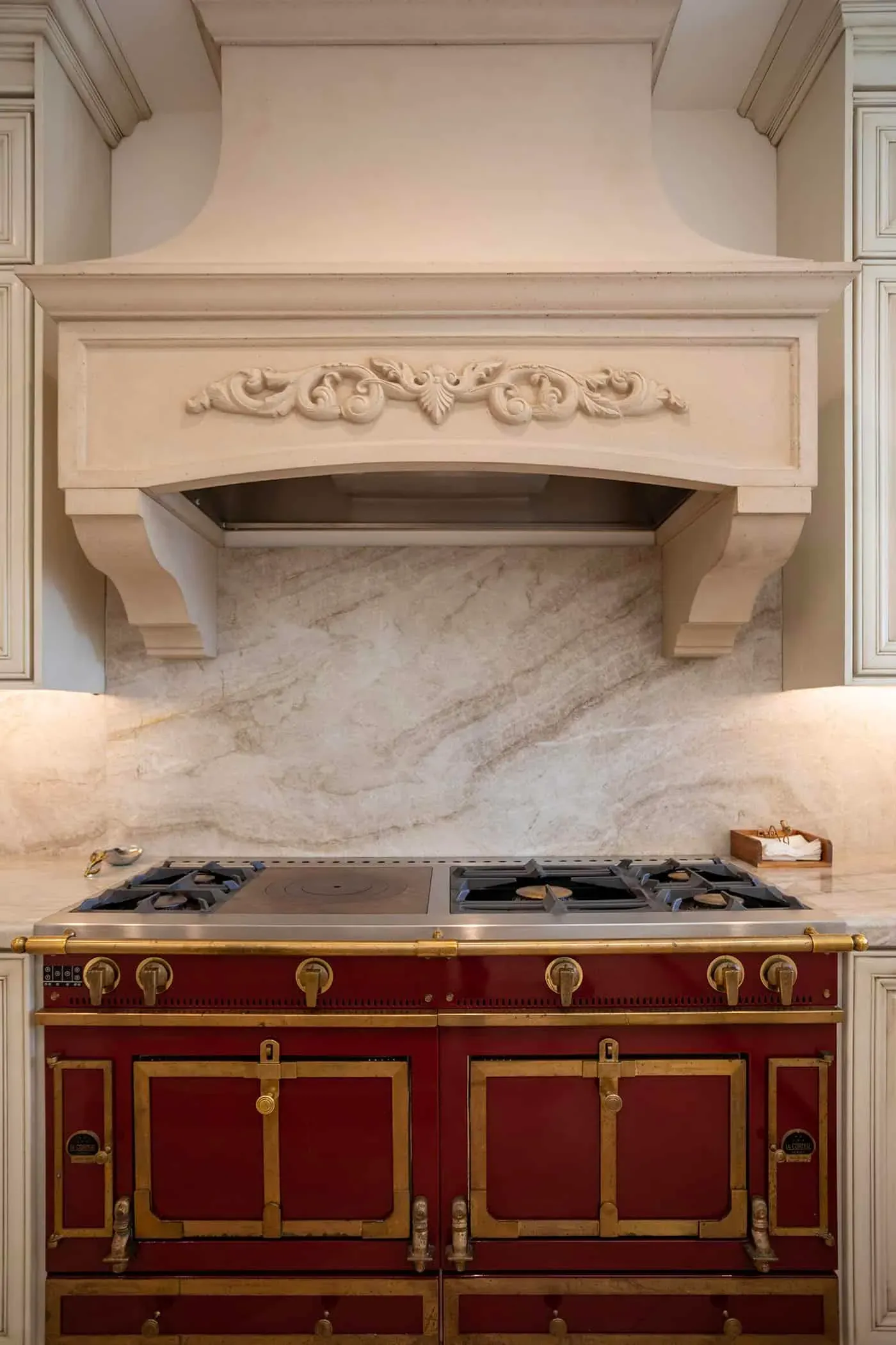 Carved hood over a red range with a book-matched stone backsplash