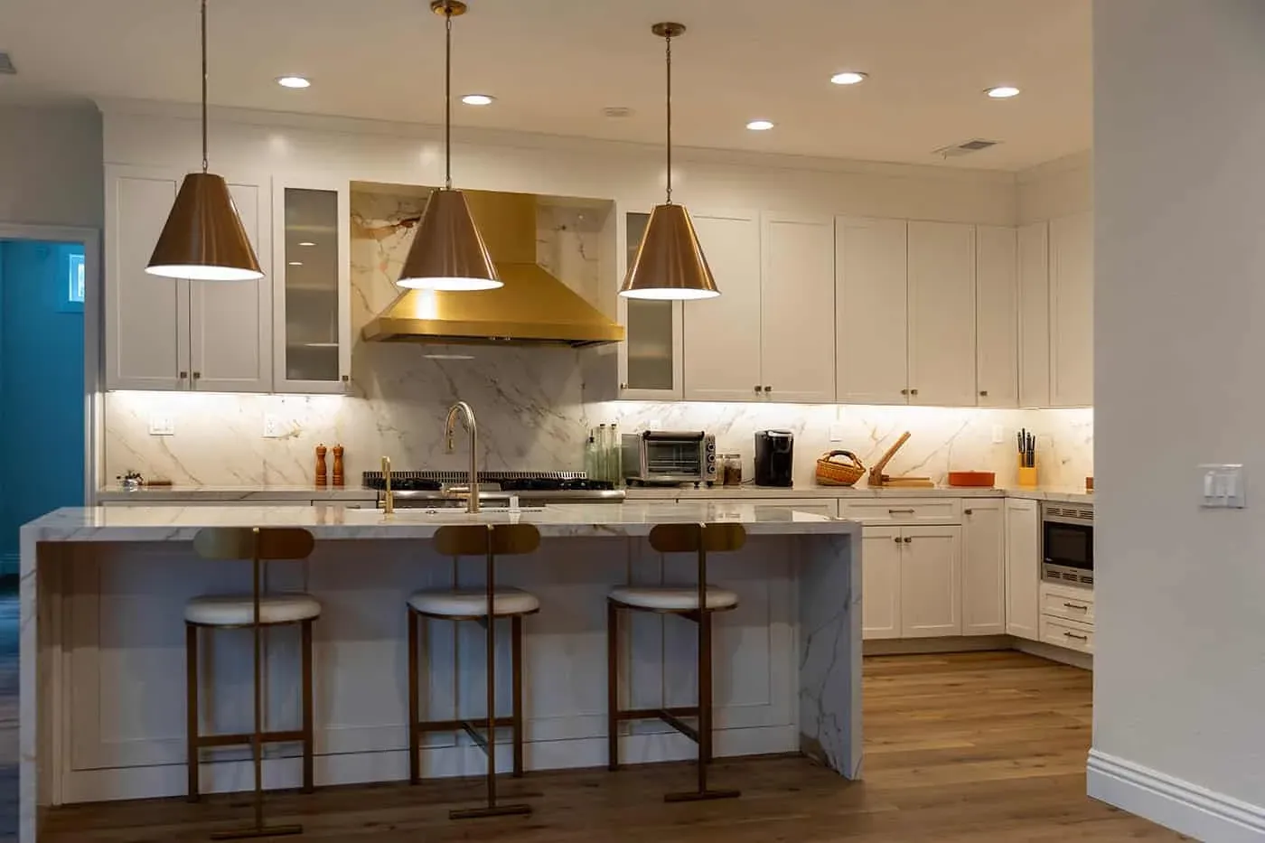 Corner view of a white kitchen with brass hood, island seating and glass-front cabinets