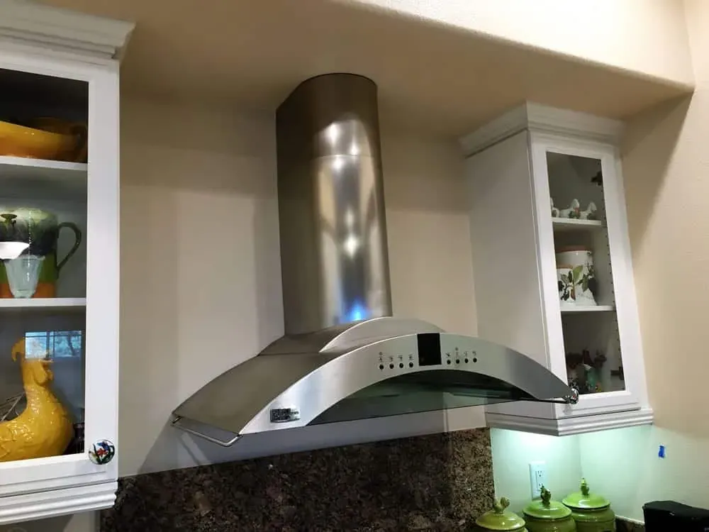 Curved stainless extractor hood over a dark granite backsplash between glass-front cabinets