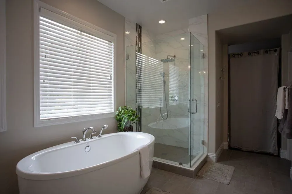 Freestanding oval tub beside a glass shower and shuttered window