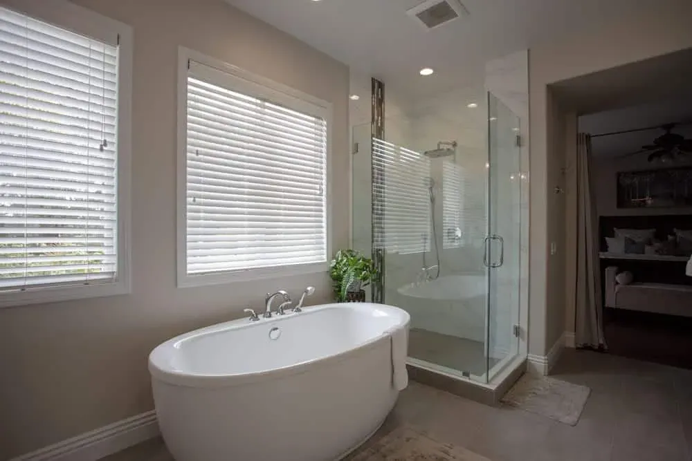 Freestanding tub beside a glass shower and shuttered windows