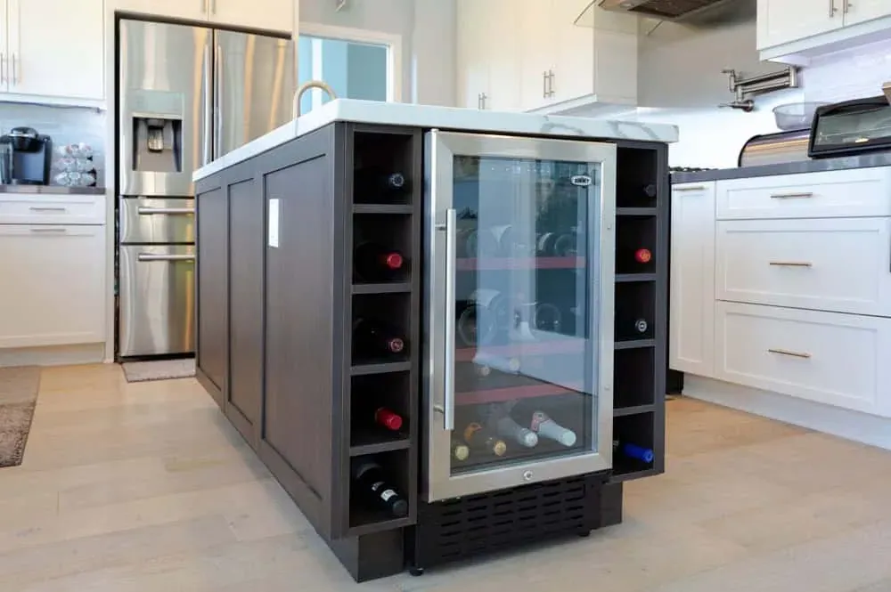 Dark kitchen island with open wine bottle cubbies and a built-in glass-front wine cooler against white perimeter cabinets