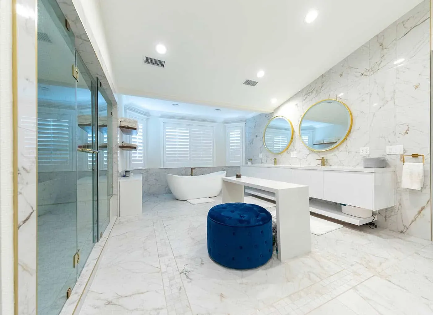 Marble bathroom with a glass shower, freestanding tub and a blue velvet stool