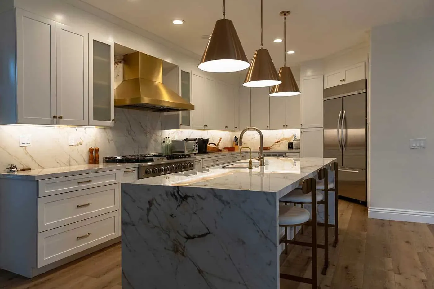 Marble island with bar stools beside a stainless refrigerator and brass hood