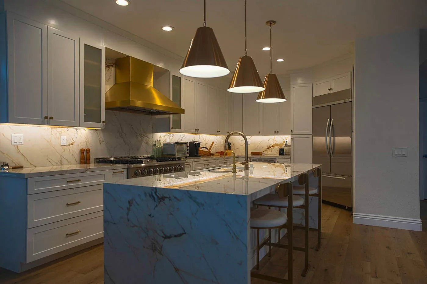 Marble island under three brass pendants with wood flooring and glass upper cabinets