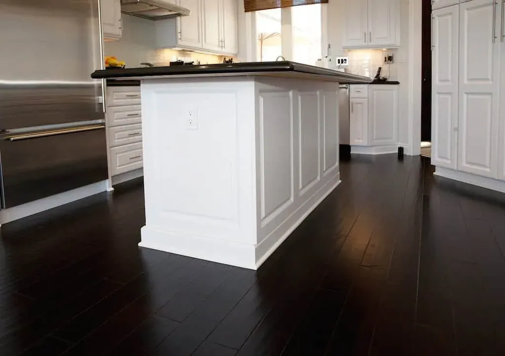 Dark stained wood plank flooring running the length of a white kitchen past a panelled island