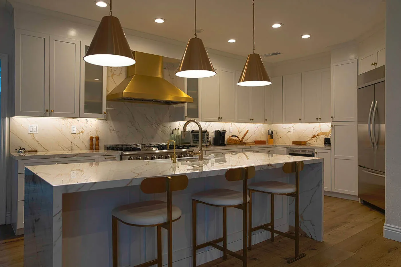 White shaker kitchen with a brass range hood, cone pendants and a marble waterfall island