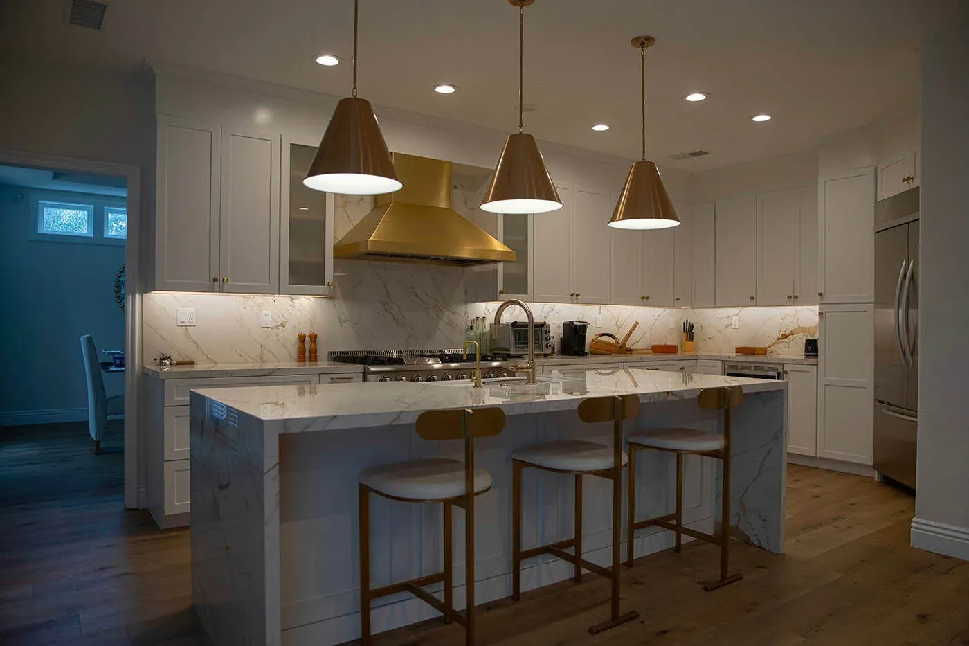 Wide view of a white kitchen with a brass hood, marble island and wood flooring